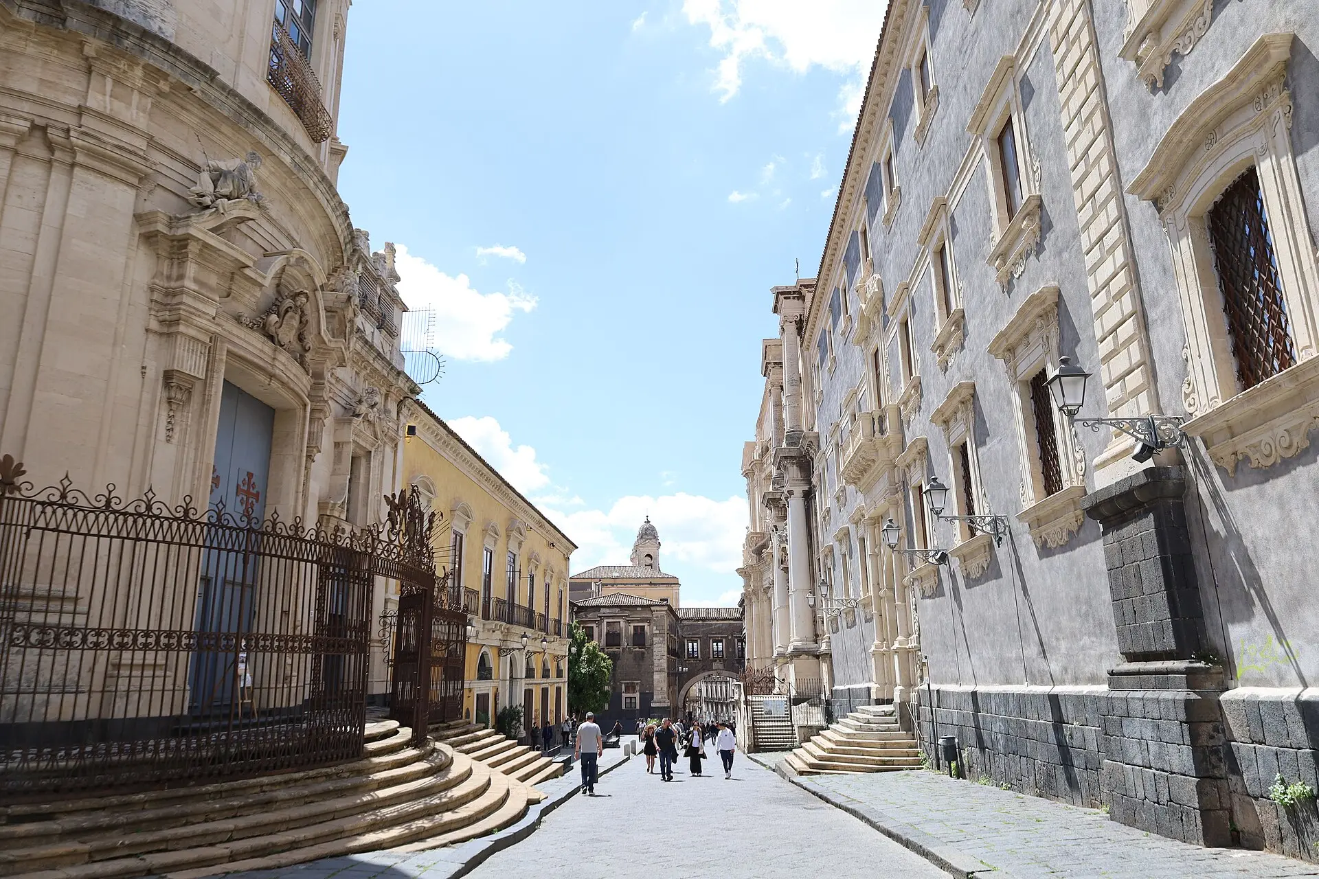 Via dei Crociferi in Catania, with the convex baroque façade and curved steps of San Giuliano on the left and the Arco di San Benedetto in the distance